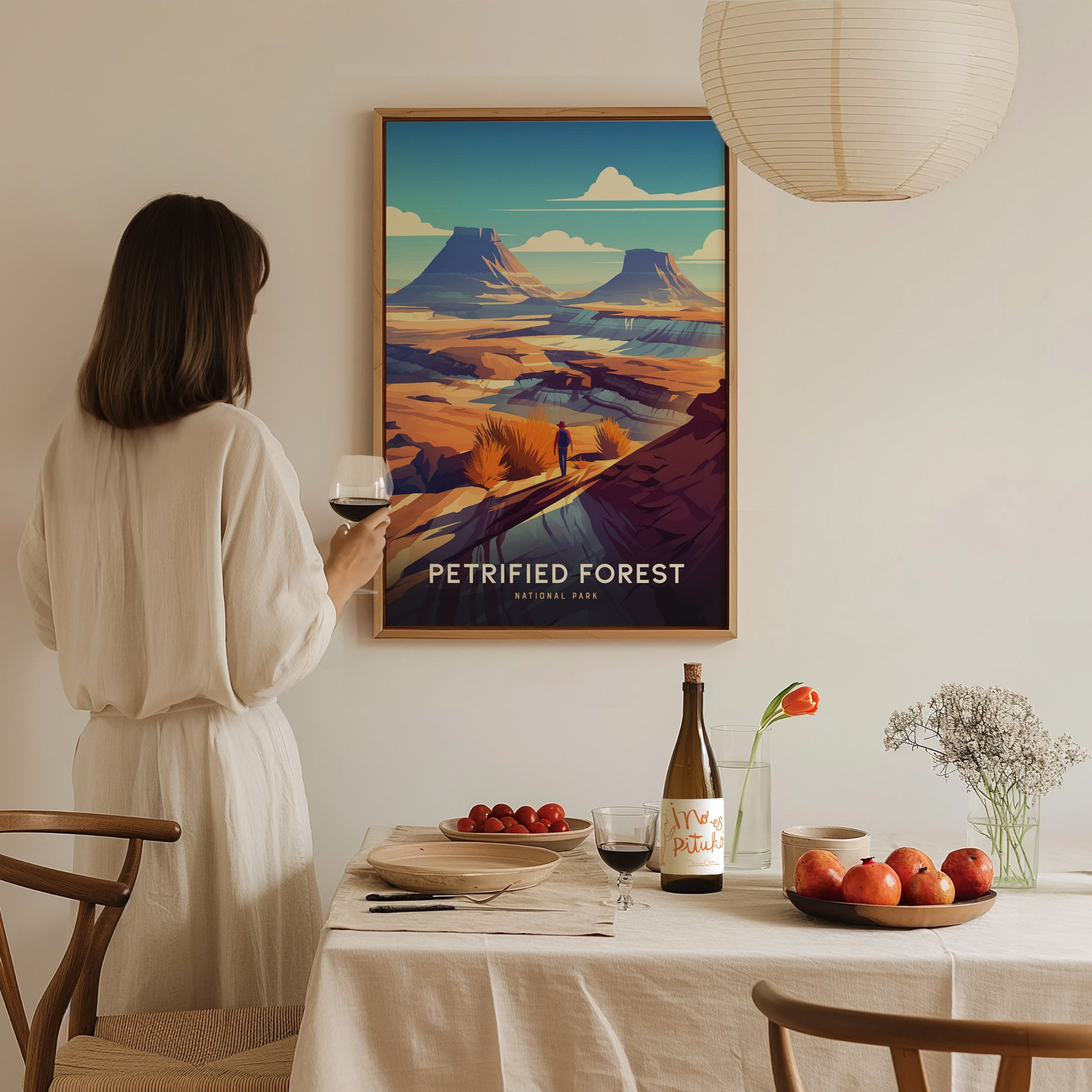A woman in a white dress admires a Petrified Forest National Park poster on a wall. A dining table with wine, apples, and flowers is in the foreground.
