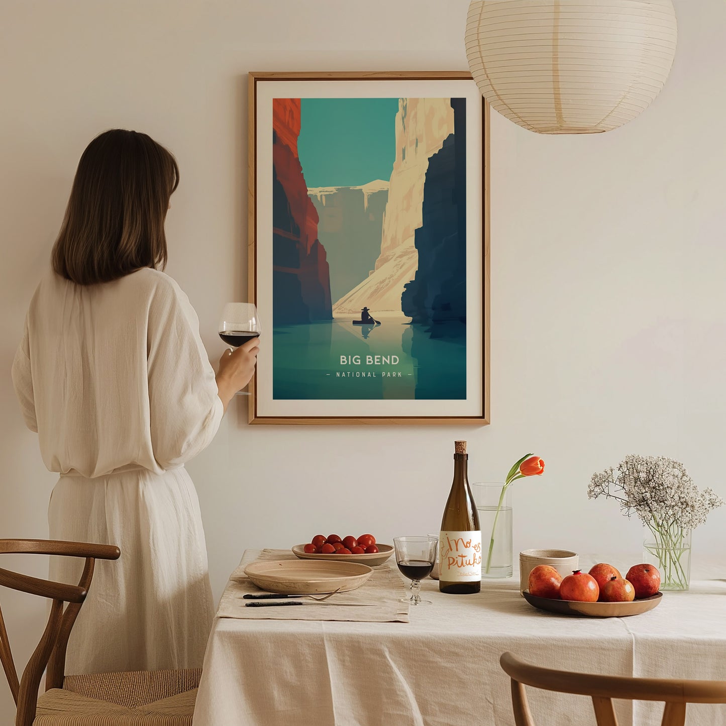 A woman in a white dress holds a glass of wine, admiring a framed Big Bend National Park poster on a wall.