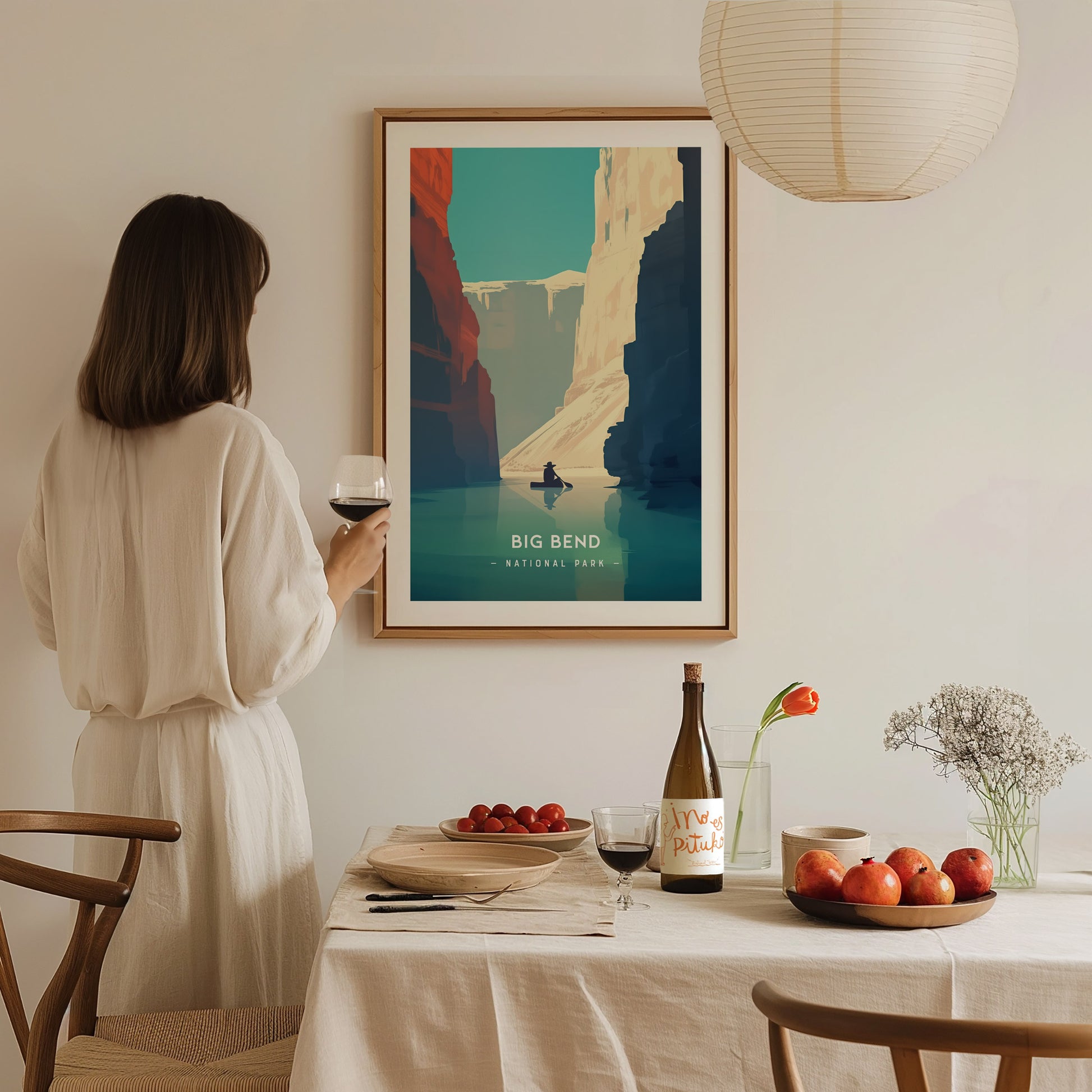 A woman in a white dress holds a glass of wine, admiring a framed Big Bend National Park poster on a wall.