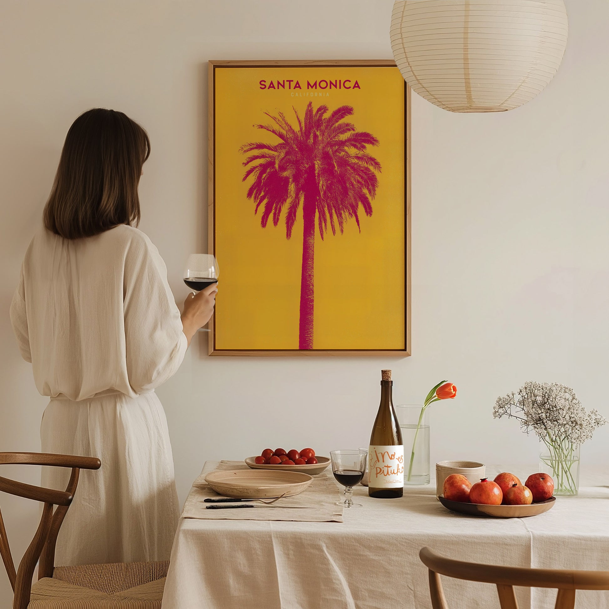 A woman in a white dress holds a glass of wine, admiring a vibrant Santa Monica poster featuring a pink palm tree.