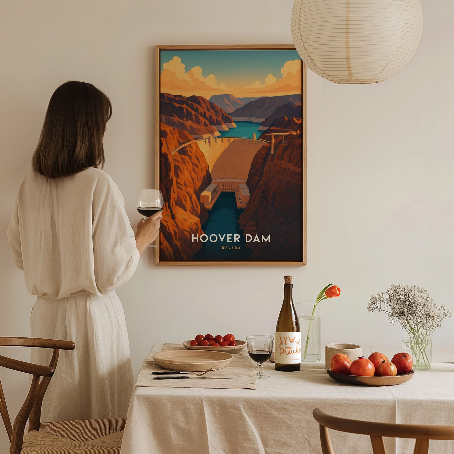 A woman in a white dress stands in a dining room, holding a glass of red wine and admiring a framed Hoover Dam poster on the wall. The table is set with fruit, a wine bottle, and a small vase with flowers.