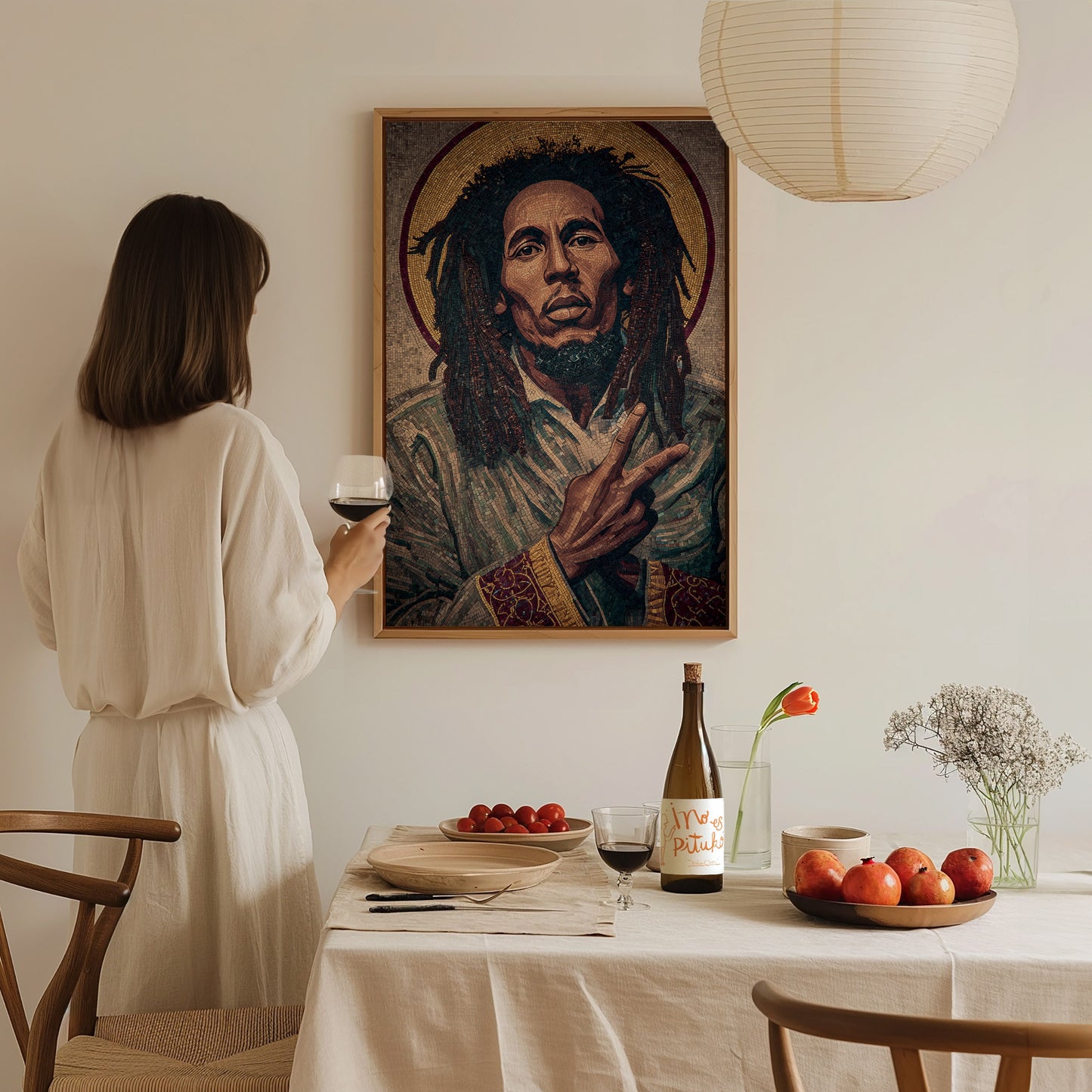 A woman in a light-colored dress stands at a dining table, holding a glass of wine and gazing at a large portrait of a man with dreadlocks on the wall.