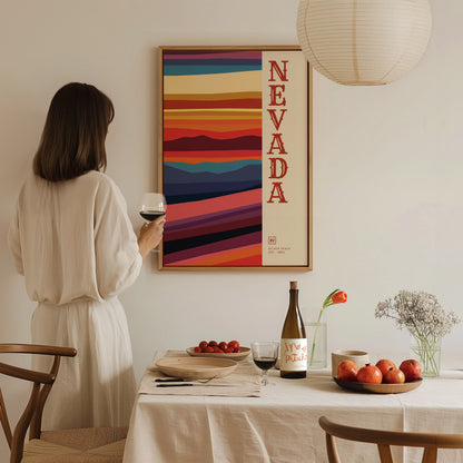 A woman in a white dress admires a colorful Nevada poster on the wall, holding a glass of wine beside a dining table with fruits and wine.