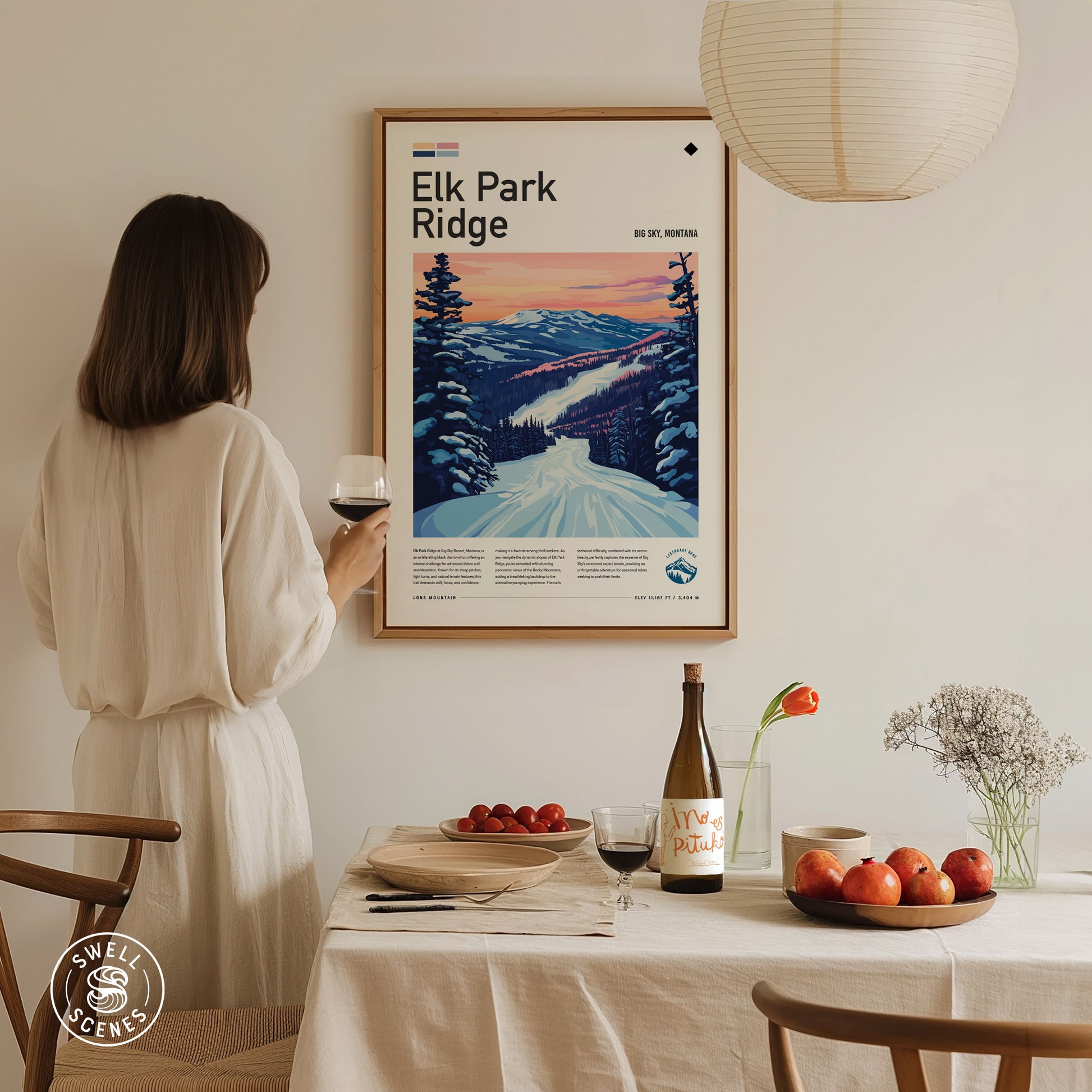 A woman in a white dress admires a framed Elk Park Ridge ski poster on a wall. A dining table is set with fruit, wine, and flowers.