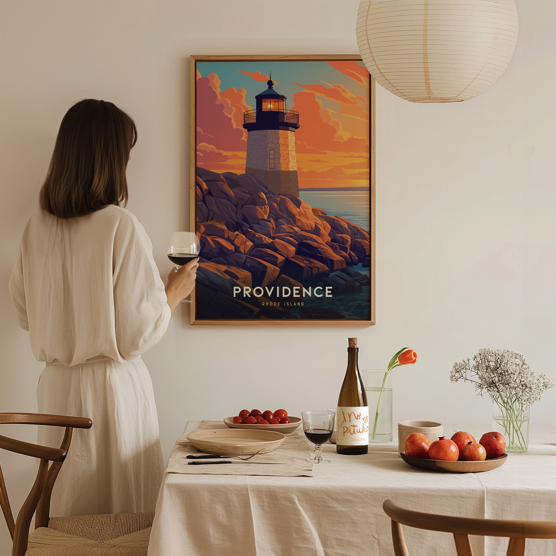 A woman in a white dress holds a glass of wine while admiring a framed poster of a lighthouse titled Providence Rhode Island.