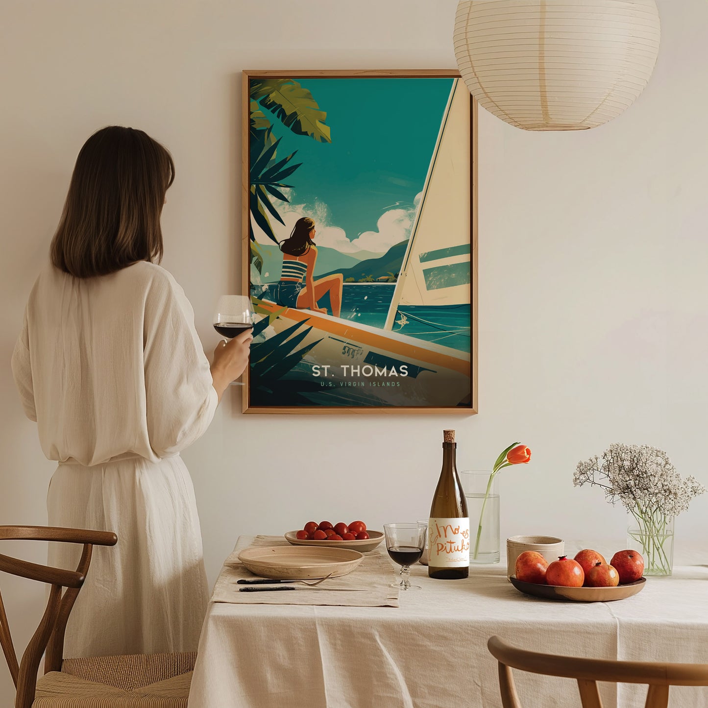 A woman in a white dress holds a glass of wine, admiring a St. Thomas poster. The table is set with fruit, a wine bottle,.