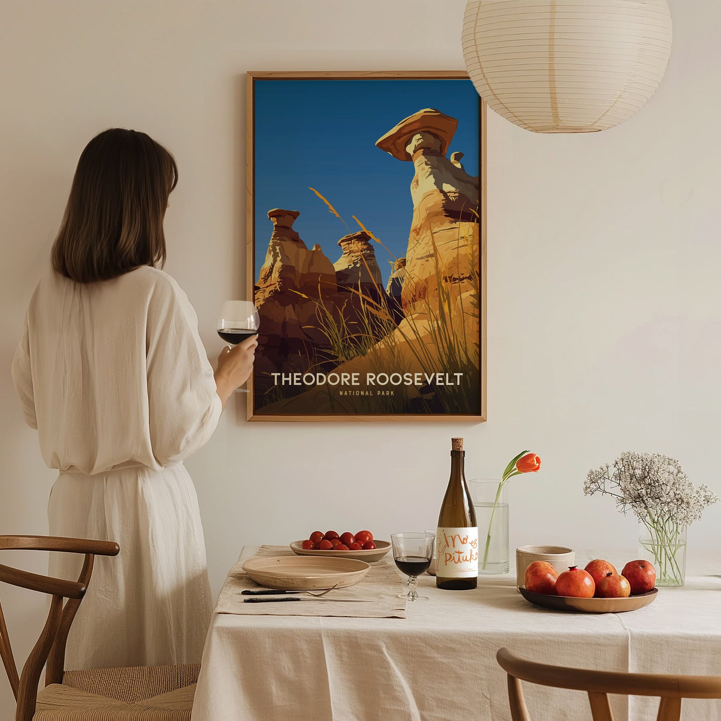 A woman in a white dress holds a glass of wine, admiring a Theodore Roosevelt National Park poster. The dining table is set with fruit, wine, and flowers.