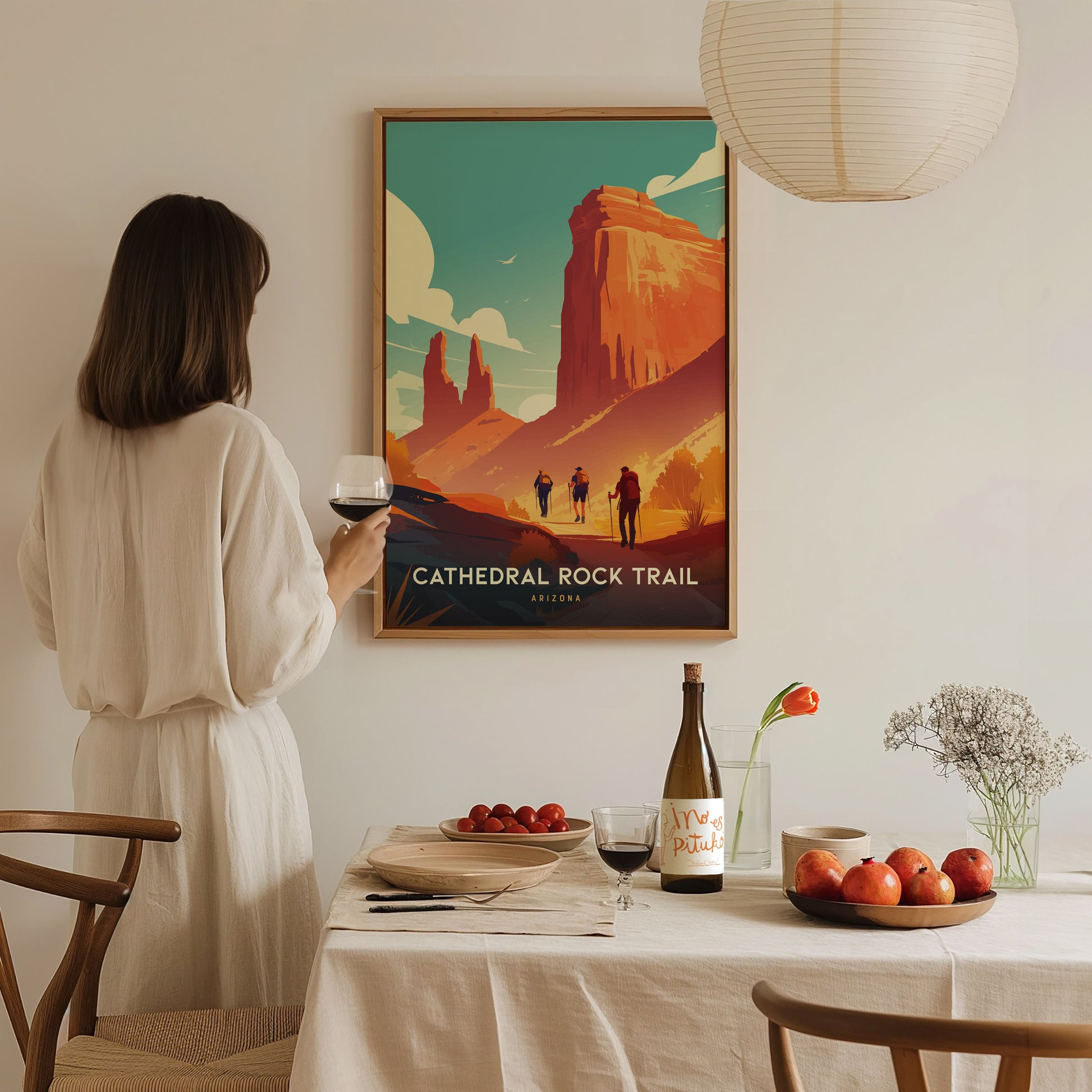 A woman in a white dress holds a glass of wine, admiring a Cathedral Rock Trail, Arizona poster. A dining table with fruits, wine, and flowers is in the foreground.