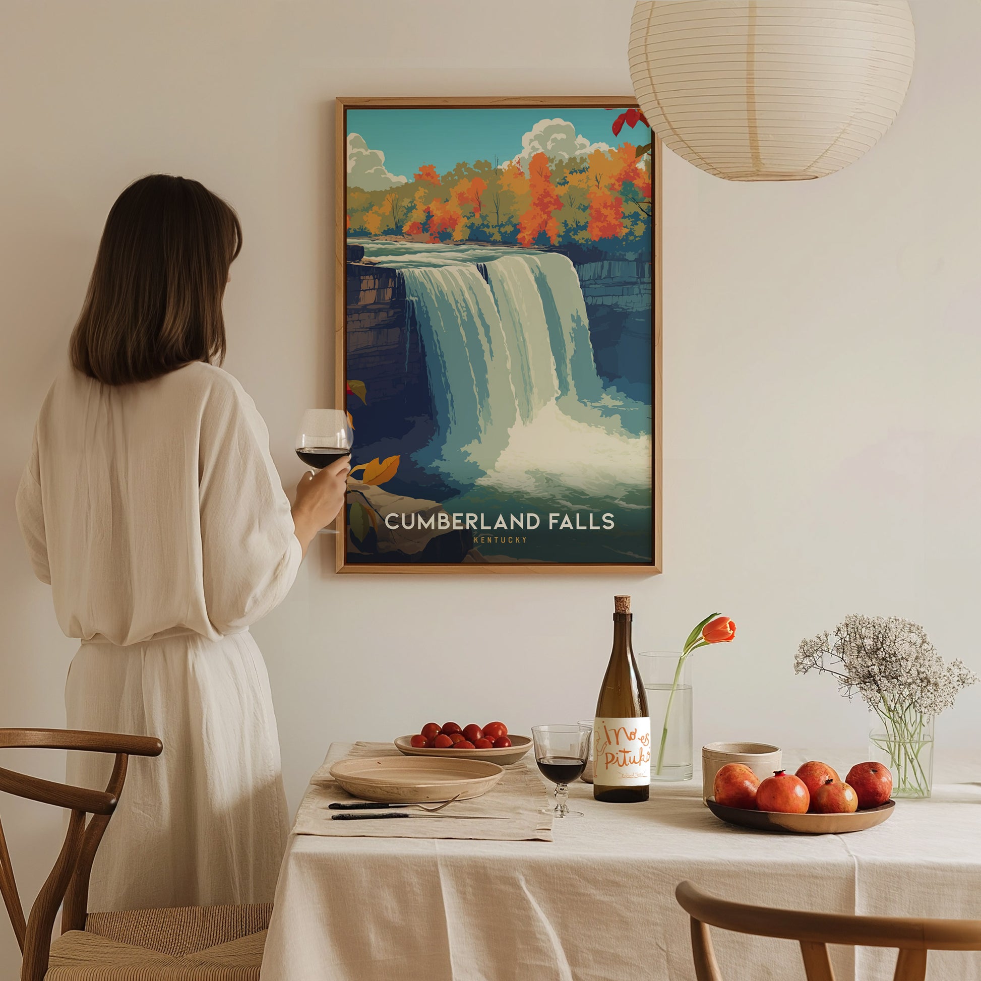 A woman in a white dress stands at a dining table, holding a glass of red wine, while admiring a framed poster of Cumberland Falls on the wall. The table is set with a bottle of wine, a plate of apples, and a small vase with flowers.