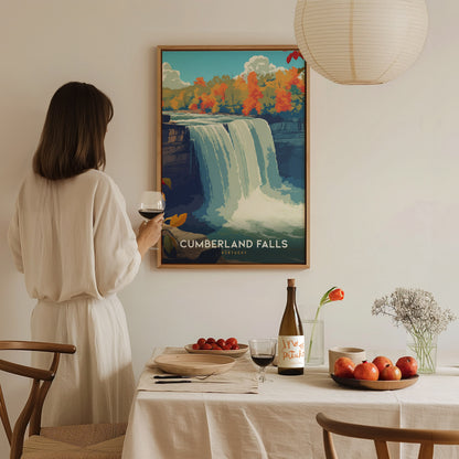 A woman in a white dress stands at a dining table, holding a glass of red wine, while admiring a framed poster of Cumberland Falls on the wall. The table is set with a bottle of wine, a plate of apples, and a small vase with flowers.