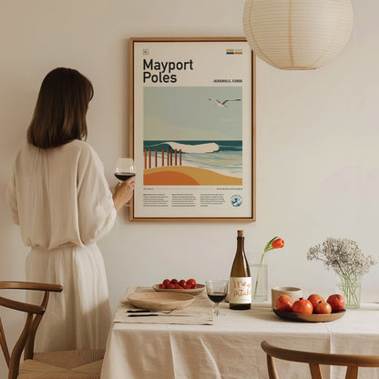A woman in a white dress holds a glass of wine, admiring a framed Mayport Poles poster on a wall above a table with fruit and flowers.