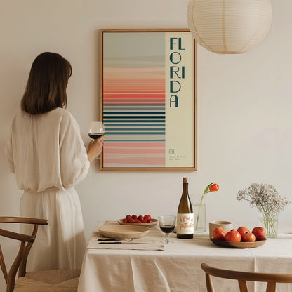 A woman in a white dress admires a framed Florida-themed minimalist poster with colorful stripes. A table set with wine, fruit, and flowers is in the foreground.