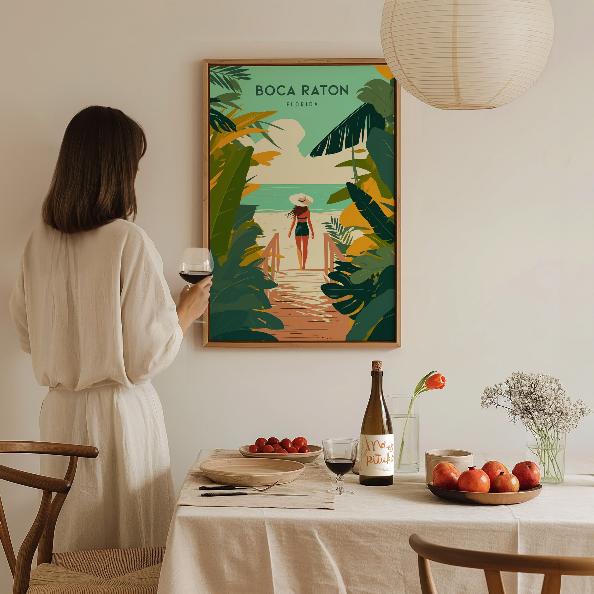 A woman in a white dress admires a Boca Raton travel print on the wall, holding a glass of wine by a table set with fruit and flowers.