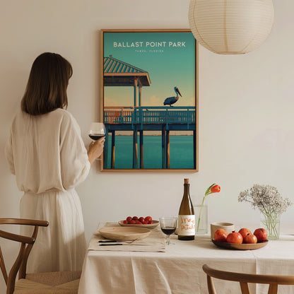 A woman in a white dress admires a Ballast Point Park poster featuring a pelican on a pier. A dining table with wine, fruit, and flowers is in the foreground.
