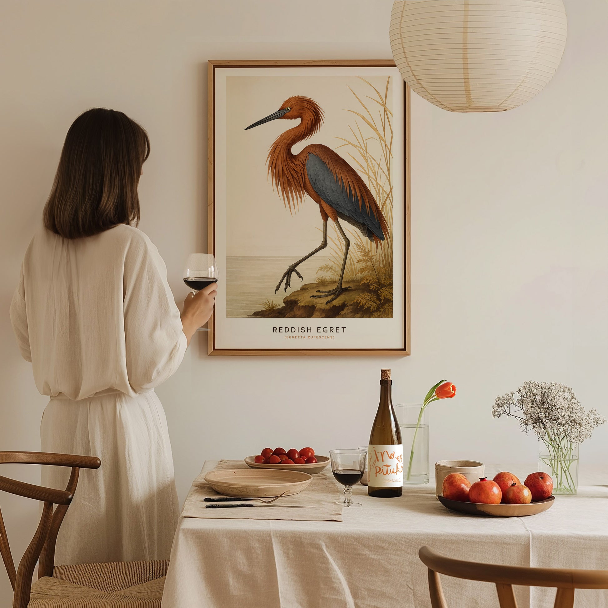 A woman in a white dress admires a framed poster of a Reddish Egret on the wall. The table below is set with wine, a bowl of apples, and a vase with flowers.