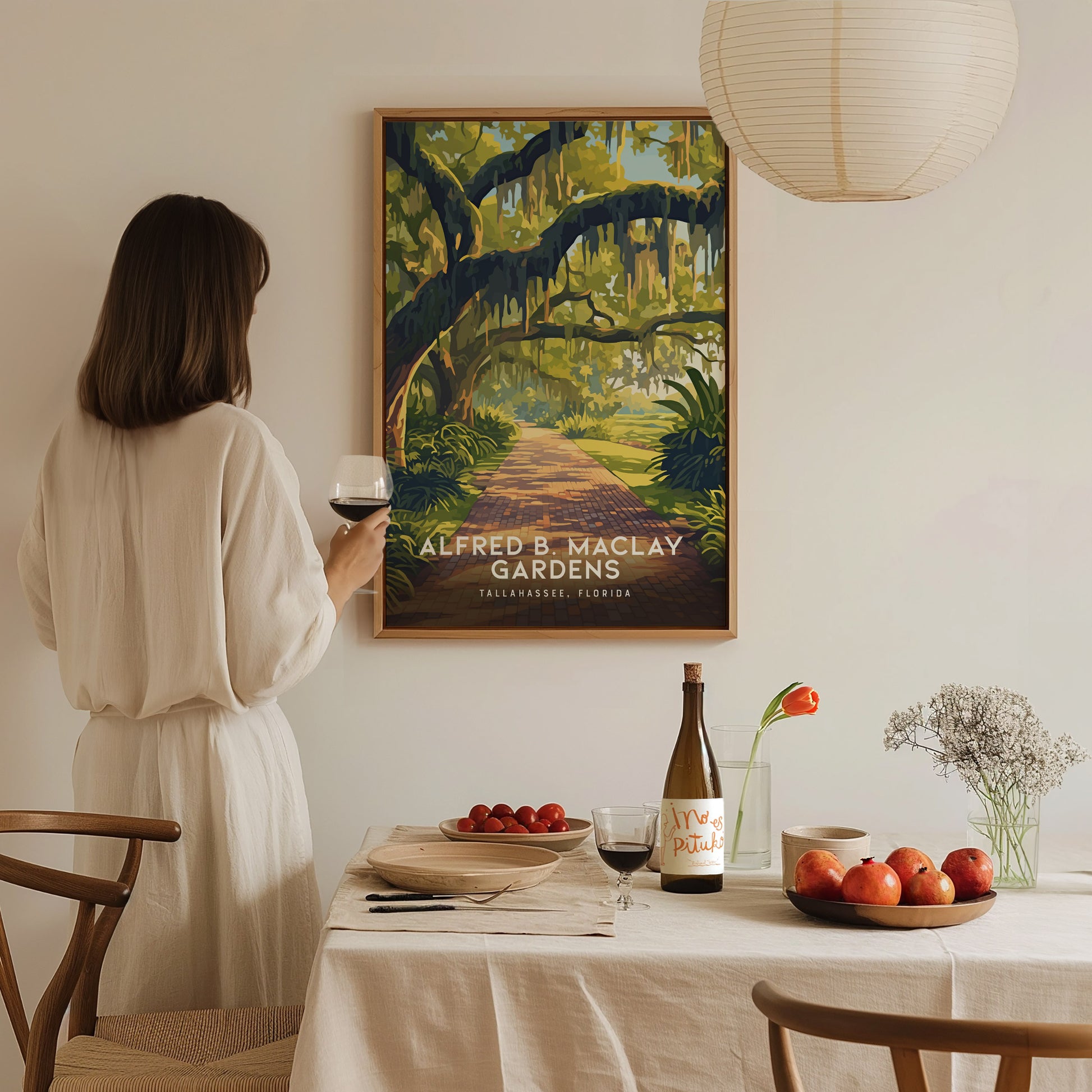 A woman in a light dress holds a glass of wine, admiring a framed Alfred B. Maclay Gardens poster. A dining table with apples and wine is in the foreground.