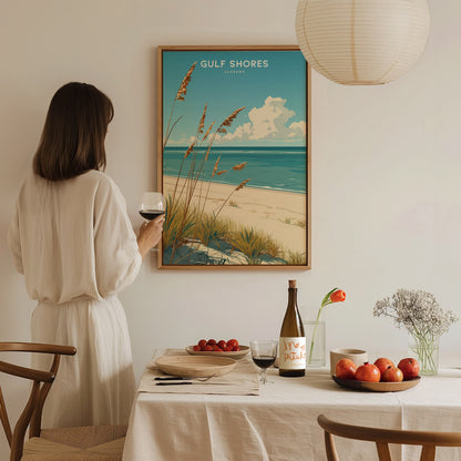 A woman in a white dress admires a Gulf Shores, Alabama beach poster on the wall, while standing beside a table set with fruit, wine, and a floral arrangement.