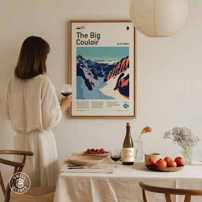 A person in a white dress holds a glass of wine, admiring a framed poster titled The Big Couloir. A table with fruit and a wine bottle is in the.