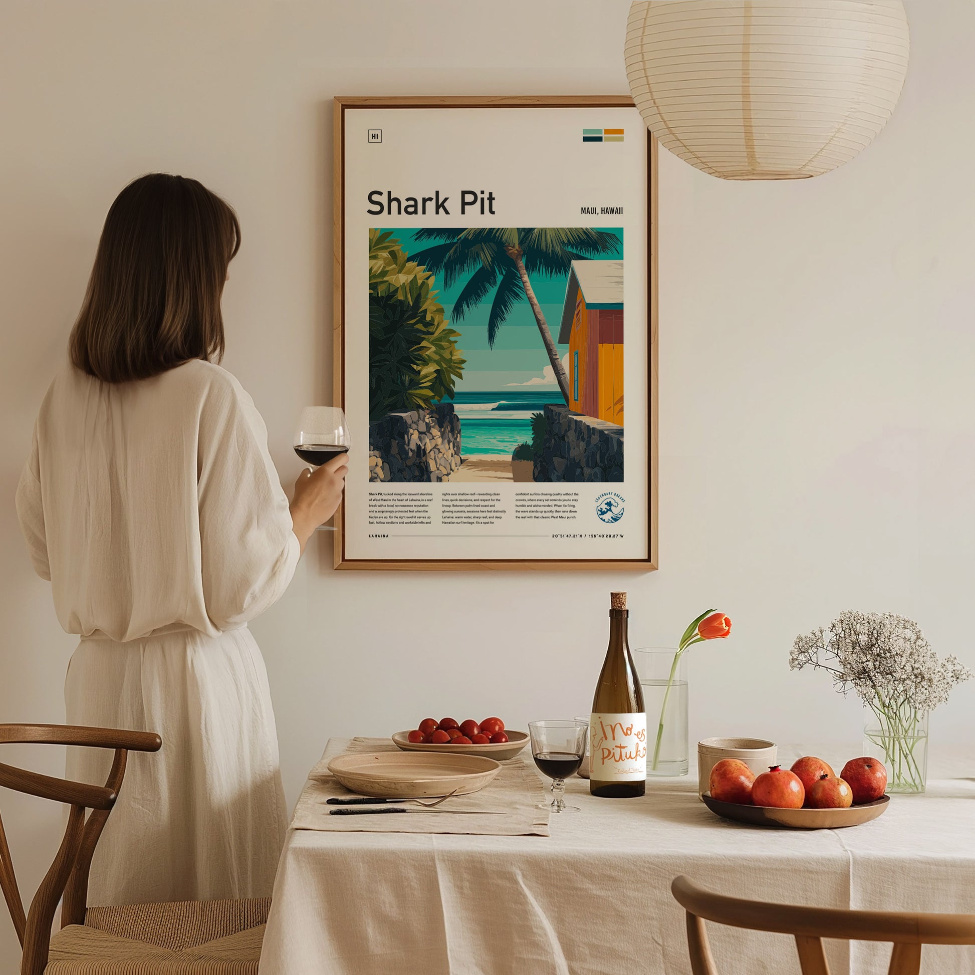 A woman in a light-colored dress stands holding a glass of wine, admiring a framed poster titled Shark Pit that features a tropical scene with palm trees and a coastal view.