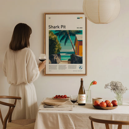 A woman in a light-colored dress stands holding a glass of wine, admiring a framed poster titled Shark Pit that features a tropical scene with palm trees and a coastal view.