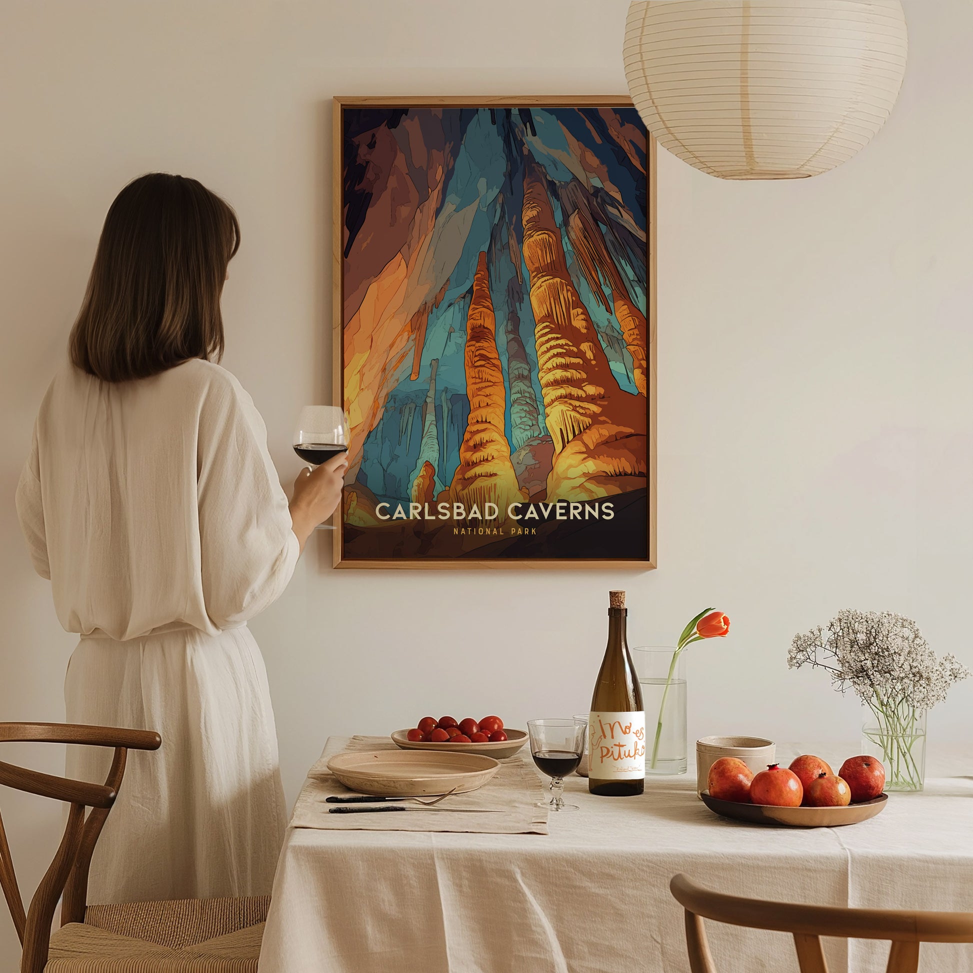 A woman in a light dress holds a wine glass, admiring a Carlsbad Caverns poster on the wall. A table with fruit, a wine bottle, and flowers is in the.