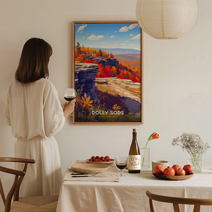 A woman in a white dress stands holding a glass of wine, gazing at a colorful poster of Dolly Sods displayed on a wall. The scene features vibrant autumn foliage, rocky cliffs, and a bright blue sky, enhancing the cozy dining atmosphere.