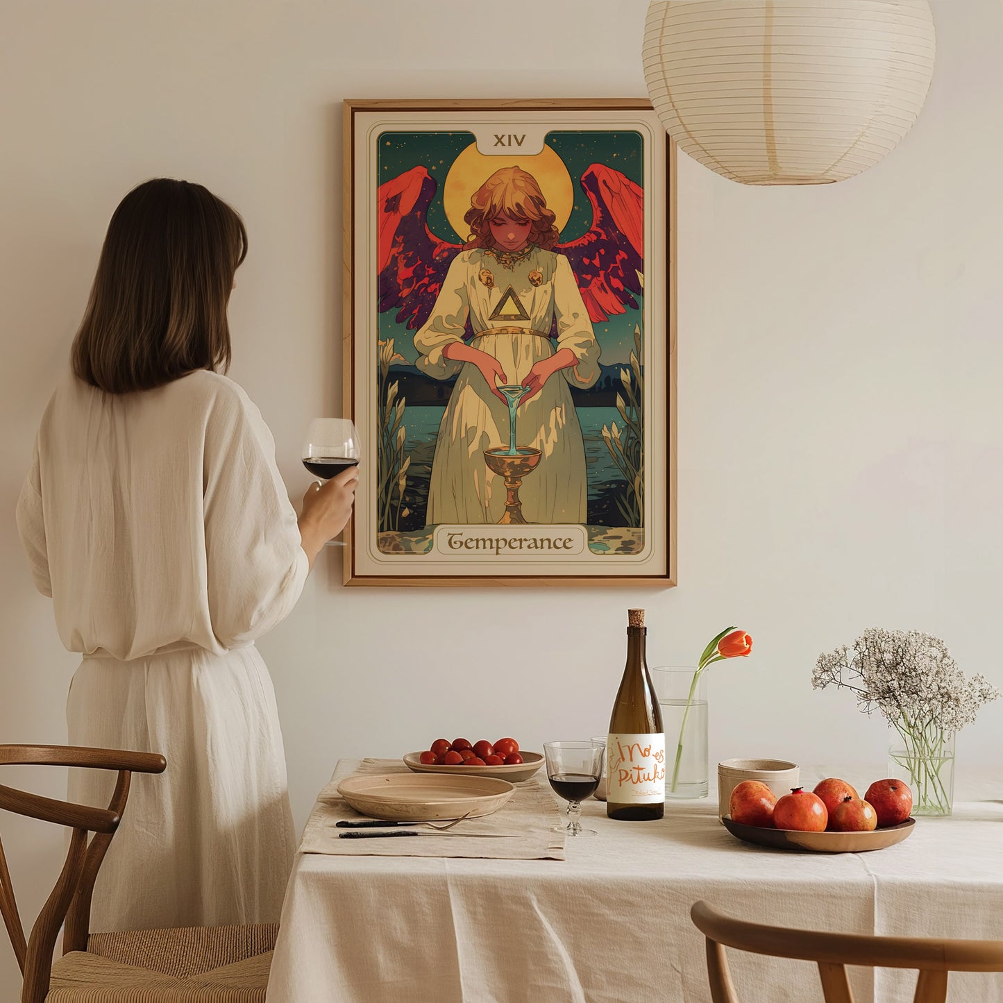 A woman in a cream-colored dress stands at a dining table, holding a glass of wine while gazing at a Temperance Tarot poster featuring an angelic figure with red wings pouring liquid between two cups.