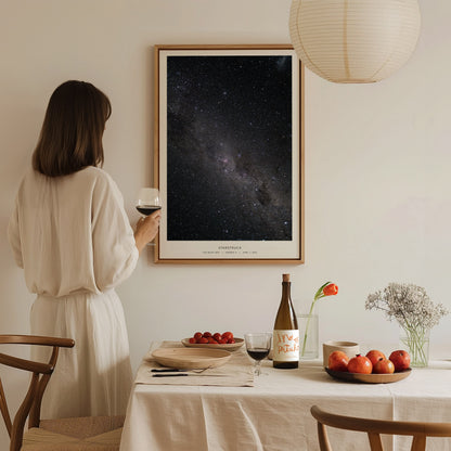 A woman in a white outfit stands in a softly lit room, holding a glass of wine and gazing at a large Milky Way night sky poster framed on the wall.