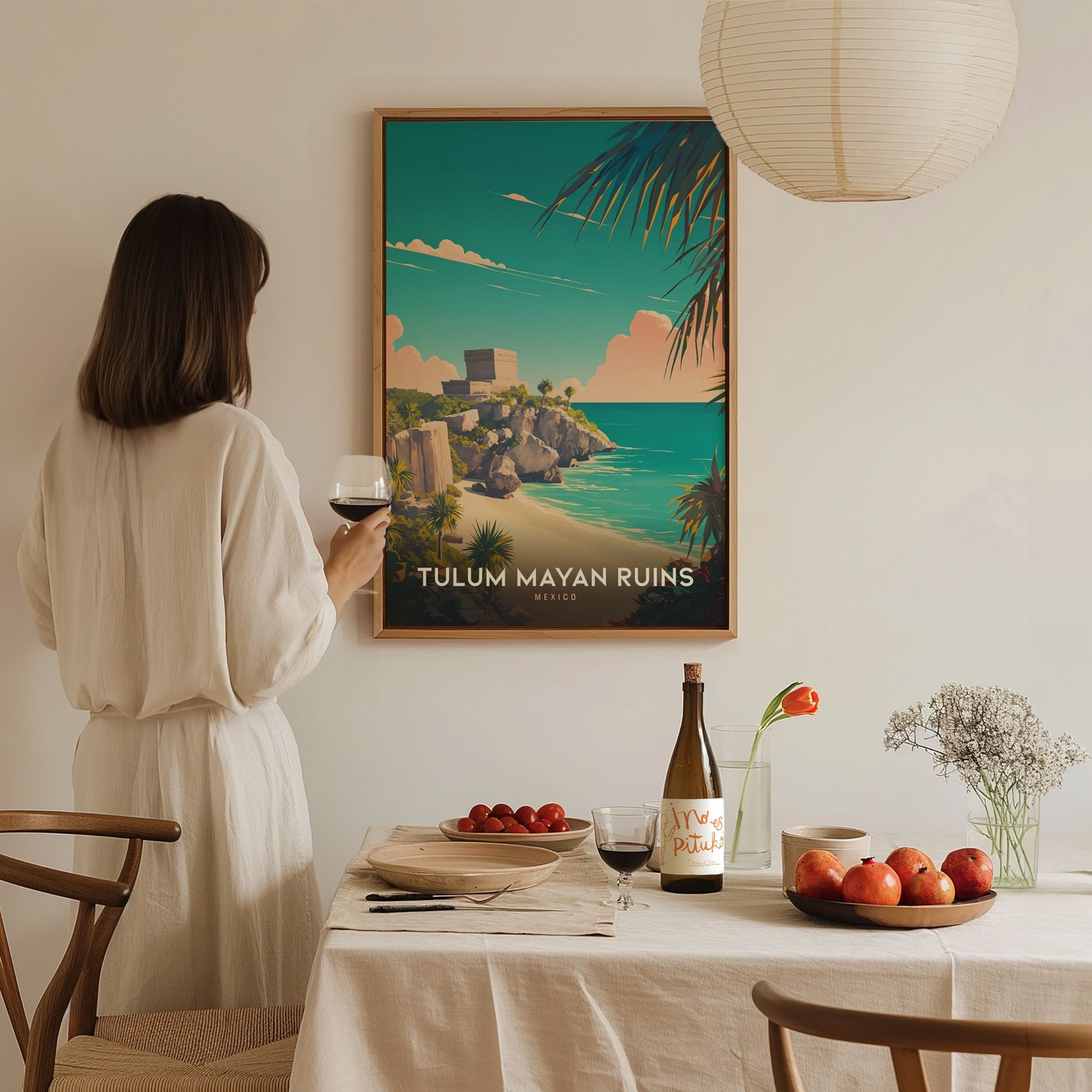 A woman in a white dress admires a framed Tulum Mayan Ruins poster on the wall, with a dining table set with fruit, wine, and flowers.