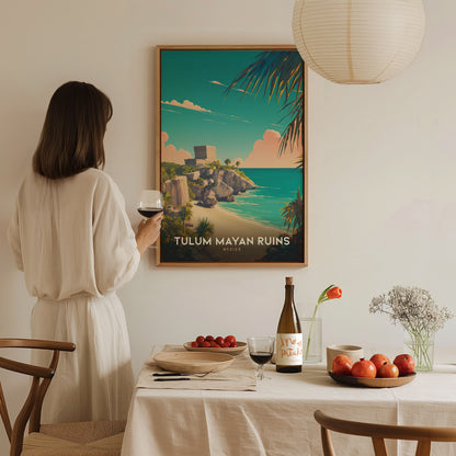 A woman in a white dress admires a framed Tulum Mayan Ruins poster on the wall, with a dining table set with fruit, wine, and flowers.