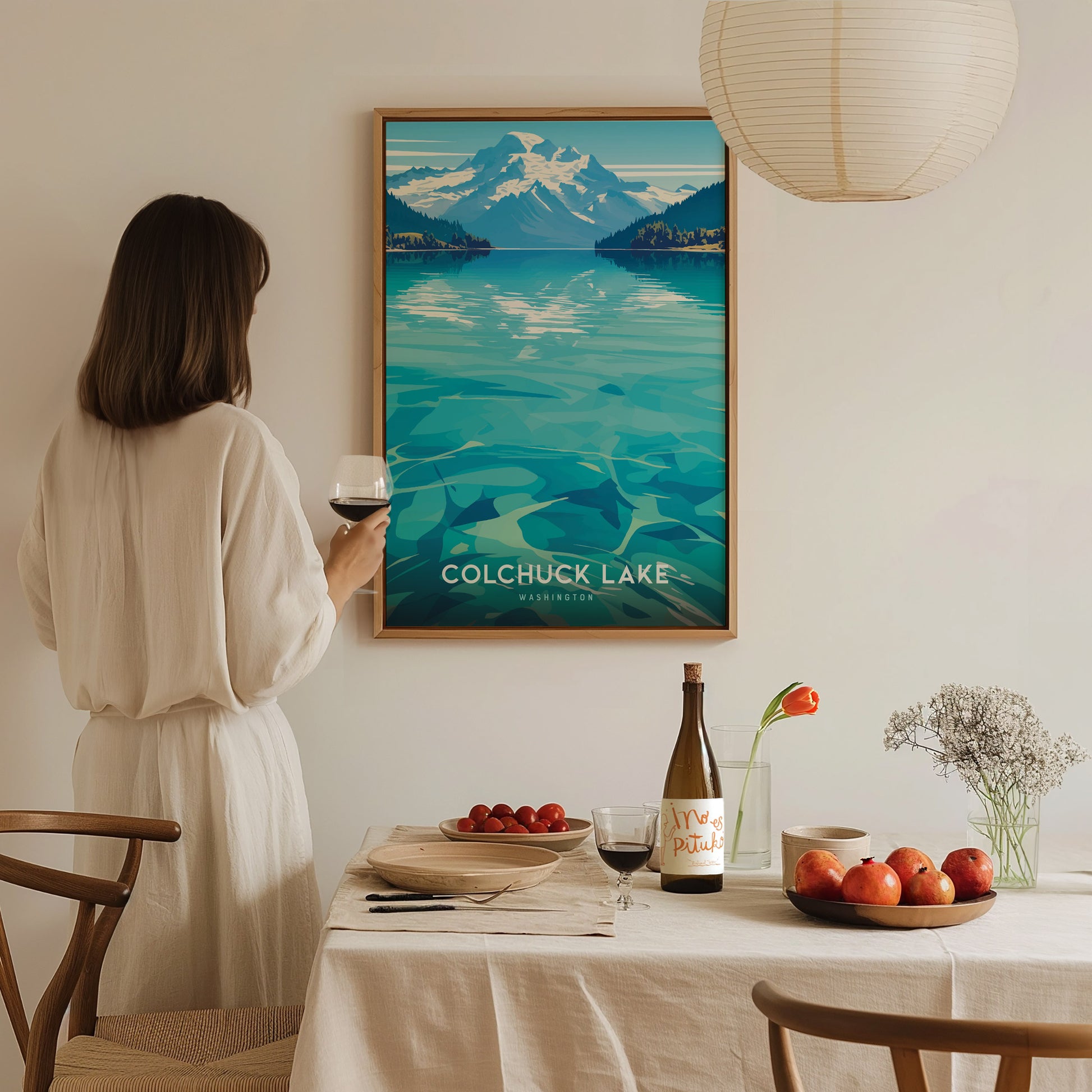 A woman in a white dress admires a Colchuck Lake poster on the wall, holding a glass of wine. A table with fruit, wine, and flowers is in the foreground.