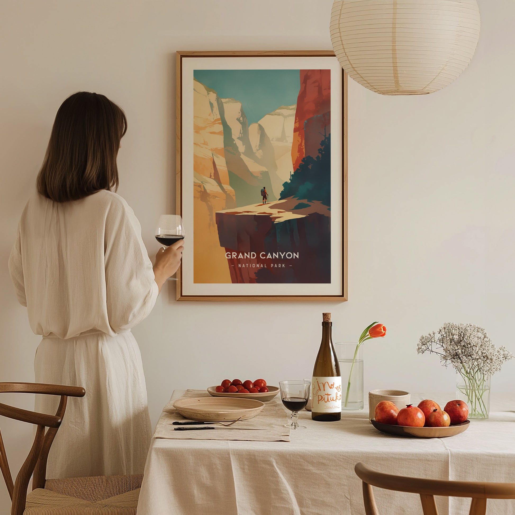A woman in a white dress holds a wine glass, admiring a framed Grand Canyon poster. The dining table features fruit, wine, and a flower vase, creating a cozy ambiance.