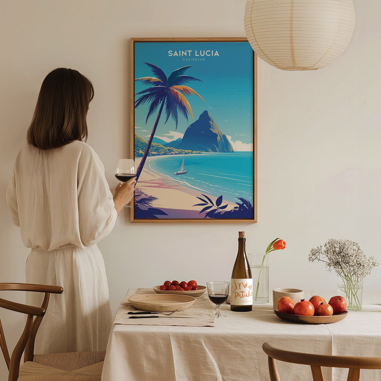 A woman in a white dress admires a Saint Lucia poster featuring a tropical beach and the Pitons. A dining table with fruit, wine, and flowers is in the foreground.