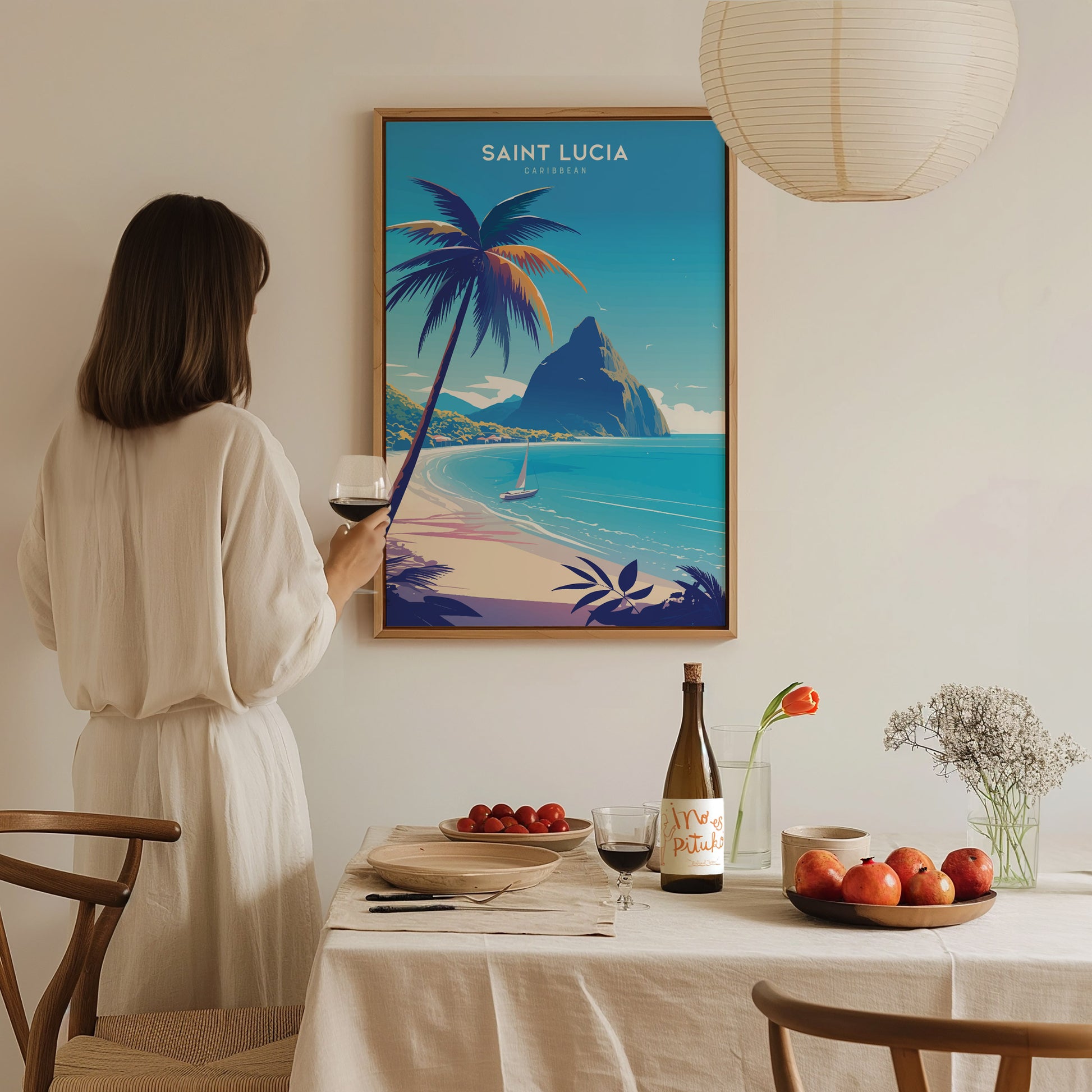 A woman in a white dress admires a Saint Lucia poster featuring a tropical beach and the Pitons. A dining table with fruit, wine, and flowers is in the foreground.