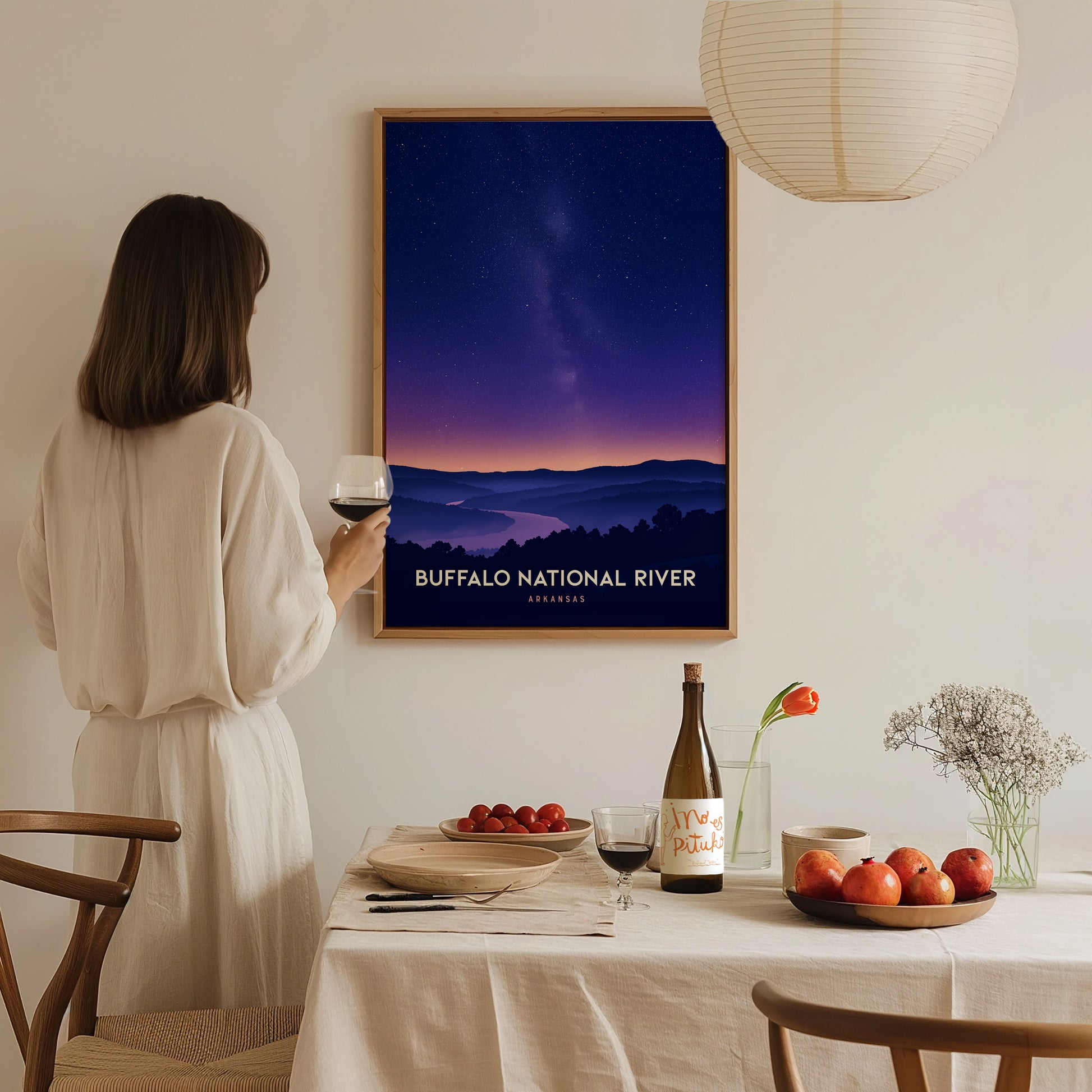 A woman in a white blouse stands at a dining table, holding a glass of wine while gazing at a framed poster of the Buffalo National River.