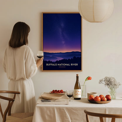 A woman in a white blouse stands at a dining table, holding a glass of wine while gazing at a framed poster of the Buffalo National River.