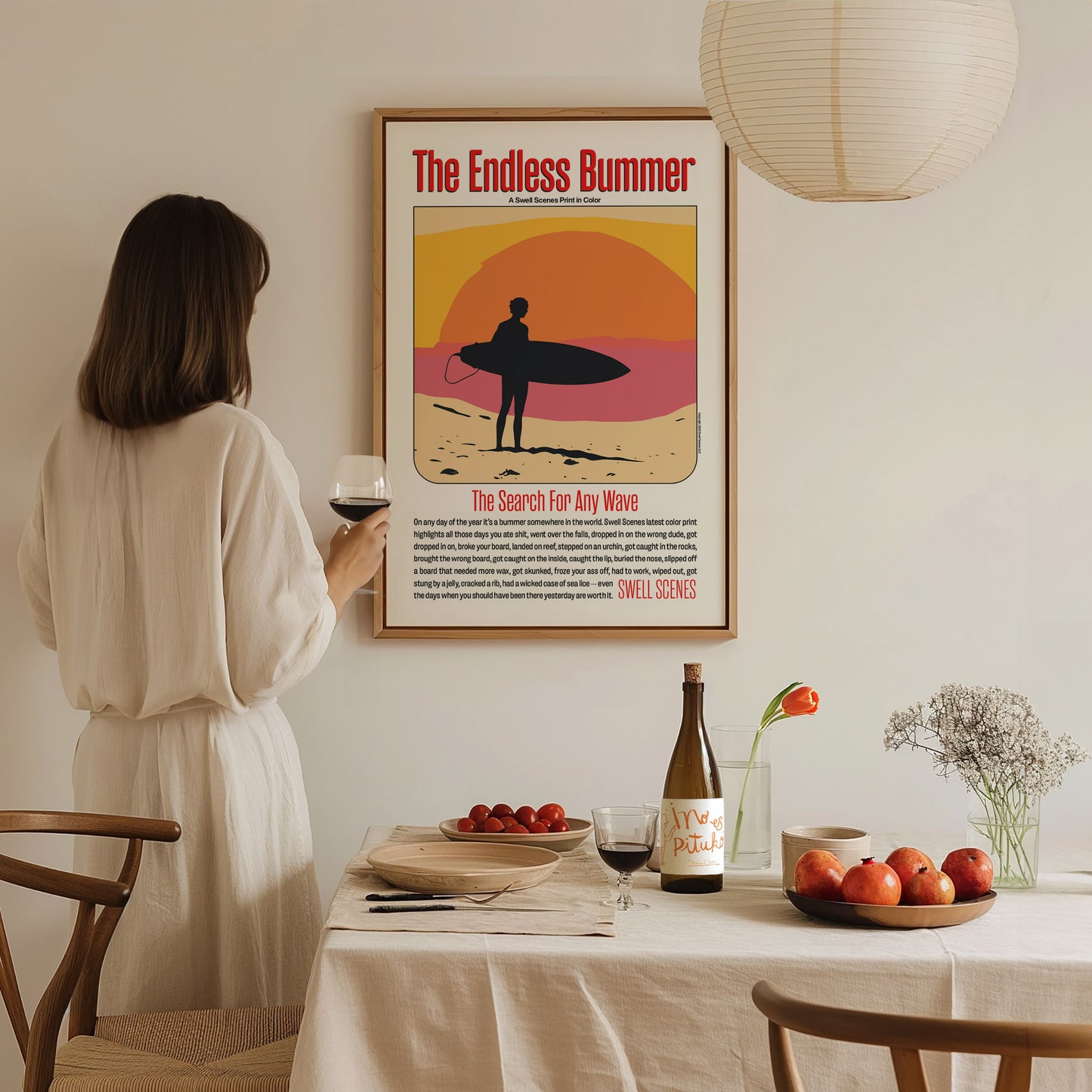 A woman in a white dress holds a glass of wine, gazing at a framed The Endless Bummer poster on the wall in a cozy dining room setting.