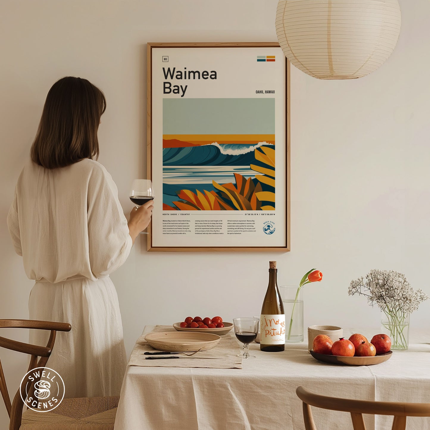 A woman in a white dress admires a colorful Waimea Bay poster on the wall. A dining table is set with a wine bottle, fruit, and a vase with flowers.