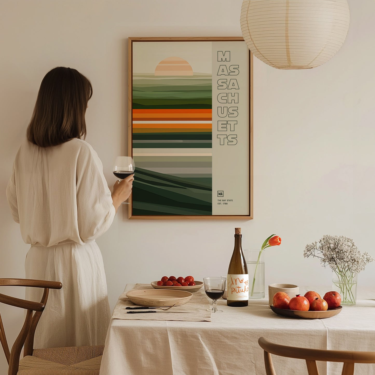 A woman in a beige dress holds a glass of wine while admiring a Massachusetts poster. The scene includes a neatly set dining table with fruits and a wine bottle.