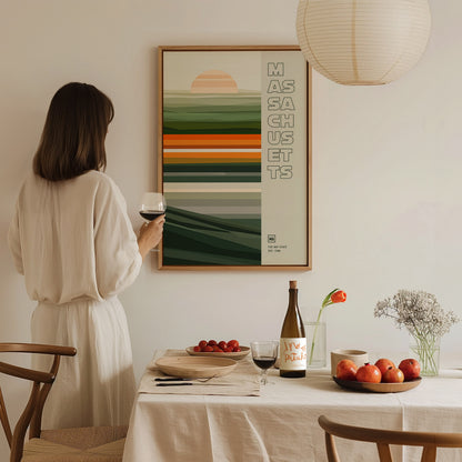 A woman in a beige dress holds a glass of wine while admiring a Massachusetts poster. The scene includes a neatly set dining table with fruits and a wine bottle.