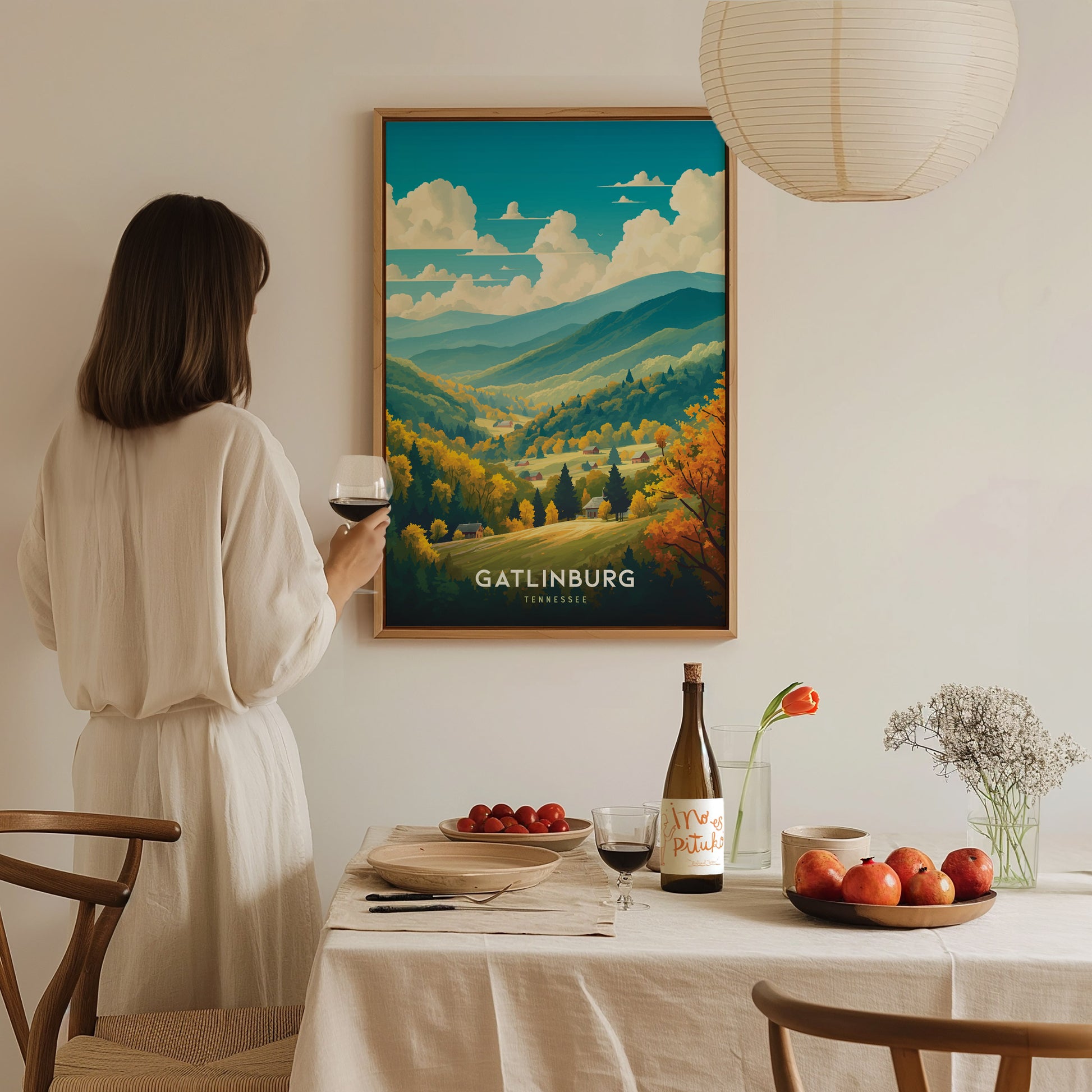 A woman in a white dress holds a wine glass, admiring a framed Gatlinburg poster on the wall. The table is elegantly set with fruit, flowers, and a wine bottle.