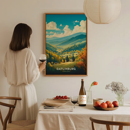 A woman in a white dress holds a wine glass, admiring a framed Gatlinburg poster on the wall. The table is elegantly set with fruit, flowers, and a wine bottle.