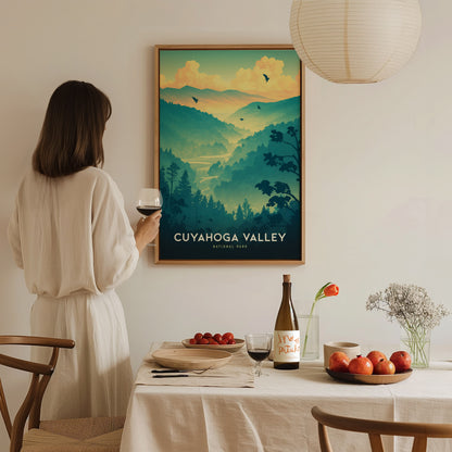 A woman in a white dress stands holding a wine glass, admiring a framed Cuyahoga Valley National Park poster. A table with fruit, wine, and flowers is in the foreground.