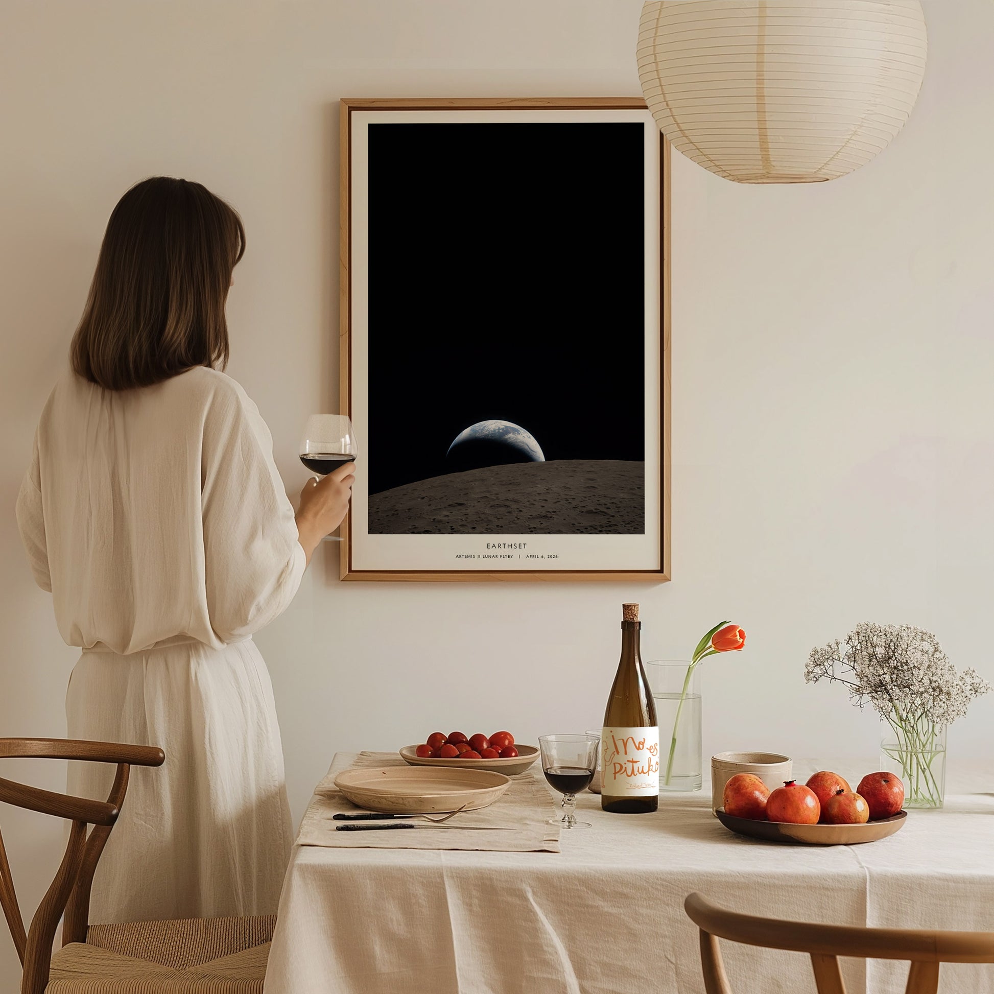 A woman in a white dress stands at a dining table, holding a glass of wine while gazing at a framed poster featuring the Earth rising above the Moon.