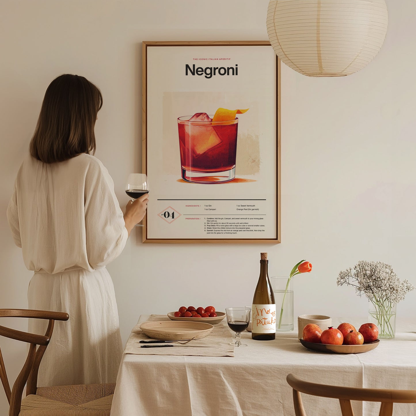 A woman in a white dress admires a framed Negroni cocktail poster while holding a glass of wine. A dining table is set with fruit, a wine bottle,.