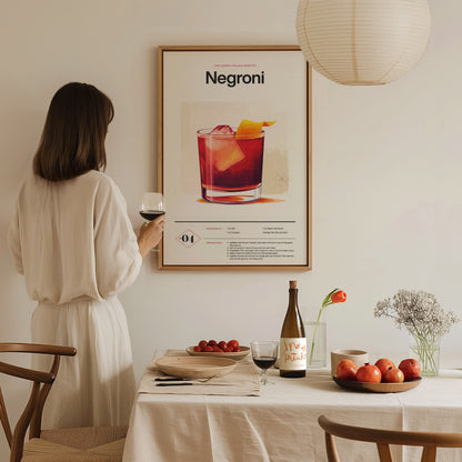 A woman in a white dress admires a framed Negroni cocktail poster while holding a glass of wine. A dining table is set with fruit, a wine bottle,.