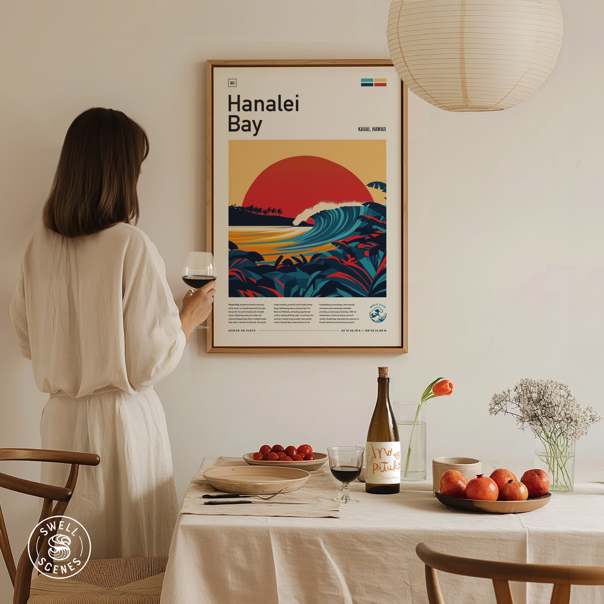 A woman in a white dress holds a wine glass, admiring a vibrant Hanalei Bay poster on the wall. A table with fruit, a wine bottle, and flowers is in.