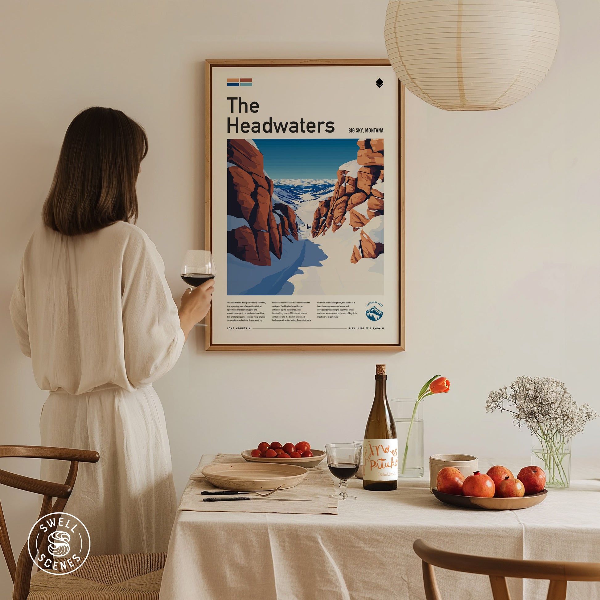 A woman in a white dress holds a glass of wine while admiring a framed poster titled The Headwaters on the wall. A table below displays fruits and wine.