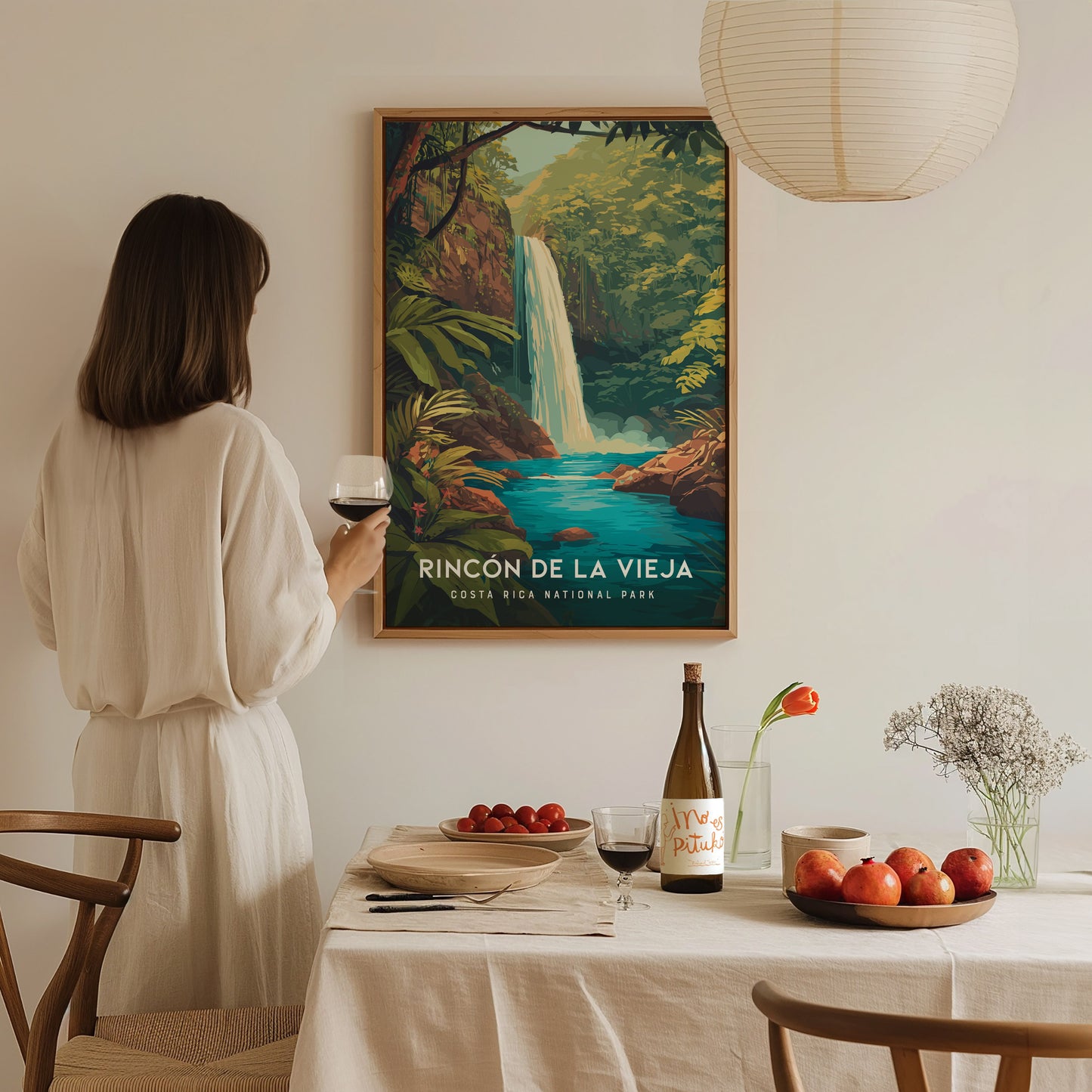A woman in a light-colored dress stands with a glass of wine in front of a dining table set with fruits and a bottle, while a vibrant poster of Rincon de La Vieja National Park featuring a waterfall hangs on the wall.