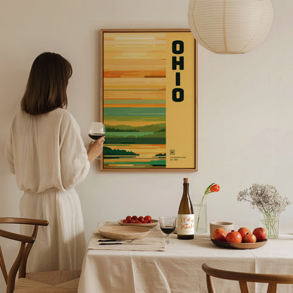 A woman in a white dress holds a glass of wine, admiring an Ohio-themed poster. The table is set with fruit, wine, and flowers, creating a cozy dining atmosphere.